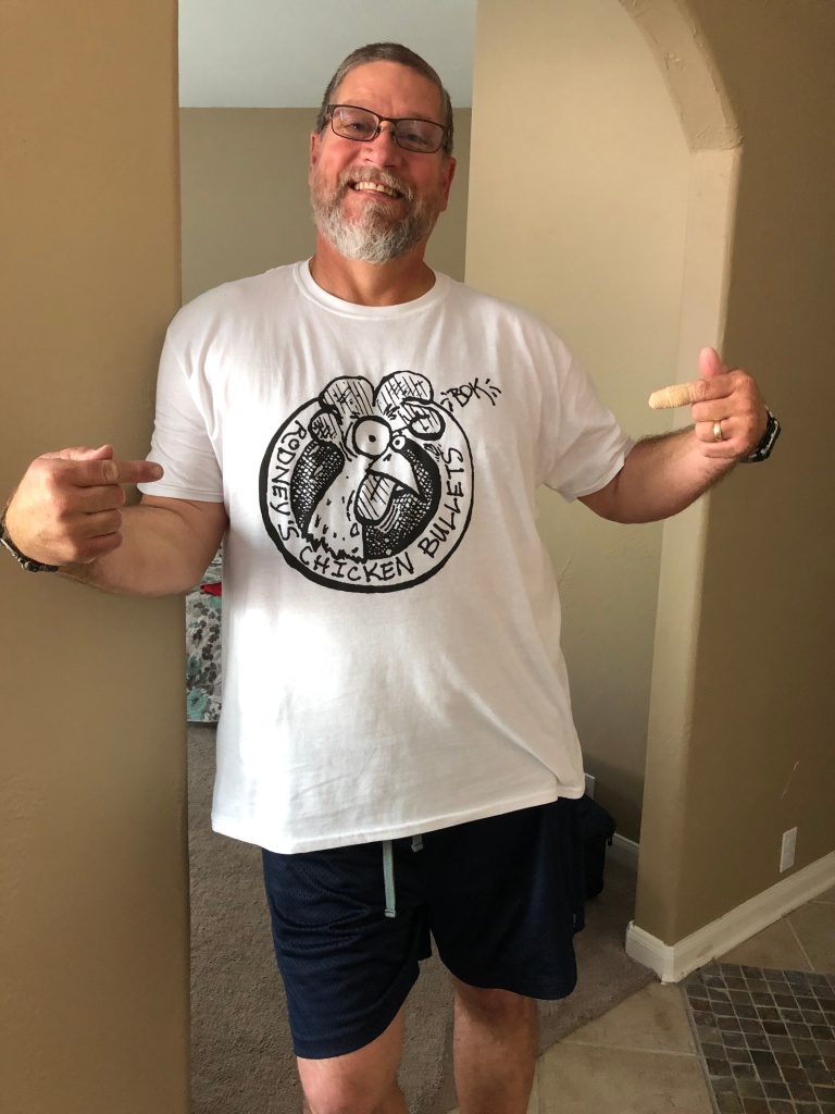 Photo of my dad wearing and pointing to a white t-shirt with an illustration of a rooster in a circle with the text "Rodney's Chicken Bullets" surrounding it.